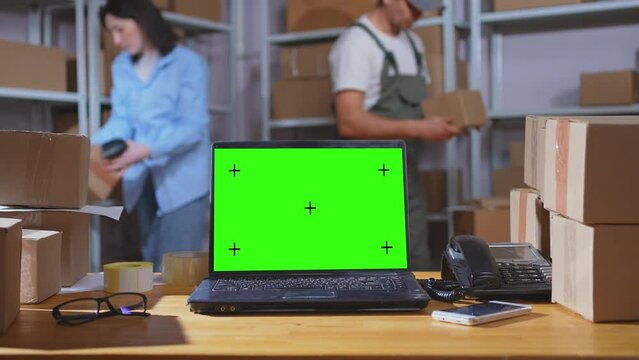 laptop with a green screen on the background of a warehouse with parcels boxes and workers