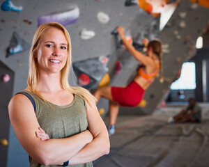 Portrait Of Female Coach In Front Of Climbing Wall At Indoor Centre With Climber In Background