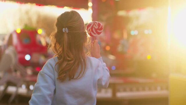 Happy smiling little girl with candy is having fun in amusement park with lights 