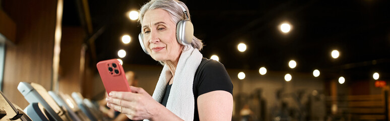 happy mature woman with towel and headphones holding smartphone and training on treadmill, banner