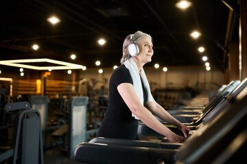 cheerful mature woman in cozy attire training on treadmill and listening music in headphones