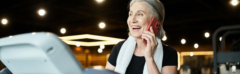 jolly mature woman with towel on shoulders talking by phone while on treadmill in gym, banner