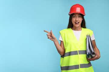 Architect in hard hat with folders pointing at something on light blue background, space for text