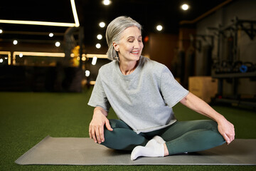 Fototapeta premium appealing mature cheerful woman in comfy sportswear sitting on floor in gym and looking away