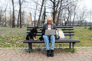 Young successful hipster businesswoman generation Z sitting on park bench working on laptop computer. Professional business female office worker in casual clothes research outside office building