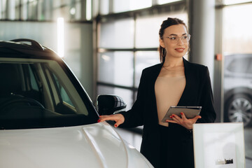 Smiling saleswoman holding tablet PC while looking at camera at new car showroom. Professional car dealer posing proudly at auto showroom, smiling to the camera, holding tablet PC.