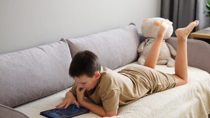 Child lies on sofa immersed in tablet game. Engrossed in digital world boy navigates through the...