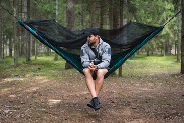 A man with a beard rests while sitting in a hammock outdoors in the forest.