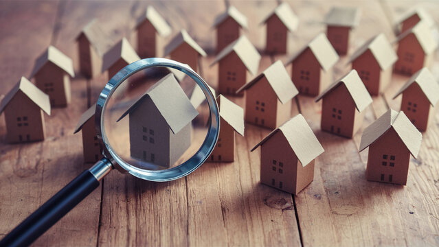 The image captures a magnifying glass focusing on a single, small house model made from cardboard among a cluster of similar miniature houses. The scene is set and situated on a wooden surface. I...