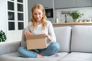 Smiling woman holding small cardboard box on knees, opening parcel