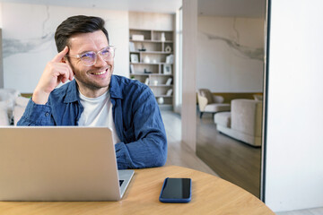 A thoughtful man with a pleasant smile works at a laptop in a tastefully decorated, airy room, suggesting creativity and focus.