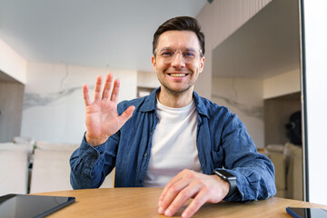 A genial man waves hello, exuding warmth and approachability amidst his home office setting,...