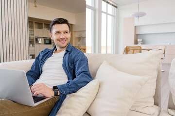 A relaxed man lounges on a sofa with a laptop, exuding ease and contentment in a well-lit, cozy living space.