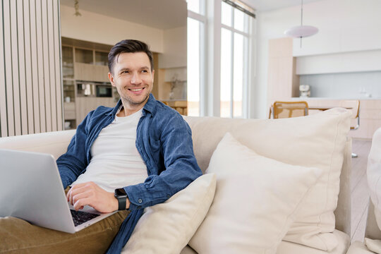 A relaxed man lounges on a sofa with a laptop, exuding ease and contentment in a well-lit, cozy living space.
