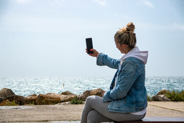 Blonde woman with bun using her mobile phone while enjoying the sea sitting on a bench on the promenade - obrazy, fototapety, plakaty