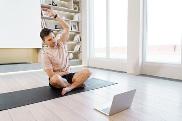 A man performs a seated side stretch in front of a laptop on a yoga mat, harmonizing physical...
