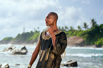 Outrageous black lgbtq person in luxury gown poses on scenic ocean beach. Non-binary ethnic fashion model in posh outfit stands gracefully at picturesque location near sea, lighthouse. Pride month.