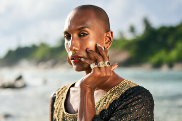 Outrageous gay black man in luxury gown, jewelry poses on scenic ocean beach. Gender fluid ethnic fashion model in posh dress, accessories looks at camera touches face with hand and rings on fingers. - obrazy, fototapety, plakaty