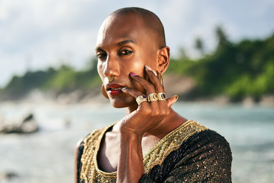 Outrageous gay black man in luxury gown, jewelry poses on scenic ocean beach. Gender fluid ethnic fashion model in posh dress, accessories looks at camera touches face with hand and rings on fingers.