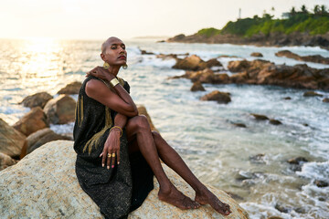 Non-binary black person in luxury dress, golden jewelry on beach rocks in ocean. Trans ethnic...