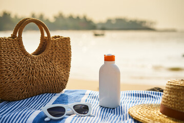 Bottle of sunscreen on striped towel next to wicker bag, straw hat, and sunglasses on sunny beach,...