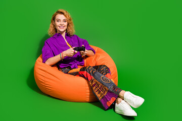 Photo portrait of lovely young lady sit beanbag hold joystick dressed stylish violet clothes isolated on green color background