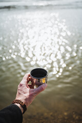 Hand with cup of coffee against the background of glare water, hiker holding glass of tea.