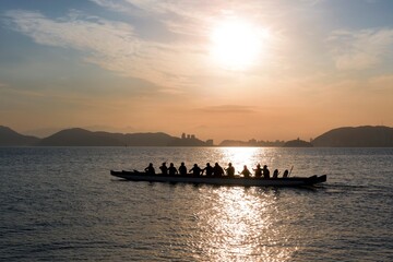 People paddling a Hawaiian canoe at sunset. In the background, the silhouette of the city's buildings. City of Santos, Brazil.