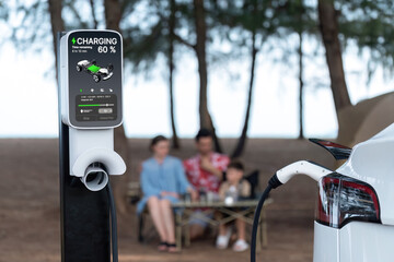 Alternative family camping trip traveling by the beach with electric car recharging battery from EV charging station with blurred family enjoying the seascape campsite background. Perpetual - obrazy, fototapety, plakaty