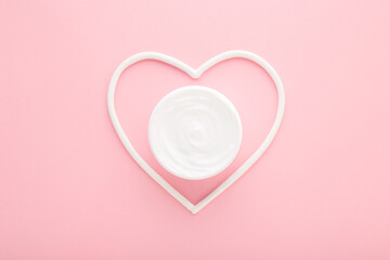 White heart shape with cream jar on light pink table background. Pastel color. Care about female face, hands, legs and body skin. Closeup. Top down view.