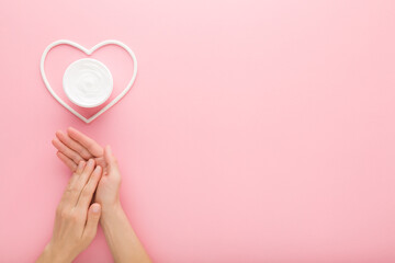 Young adult woman hands and white heart shape with cream jar on pastel pink table background. Care about clean and soft body skin. Closeup. Point of view shot. Empty place for text. Top down view.