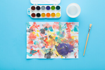 Paintbrush, watercolor palette and drawn colorful spots on white paper. Light blue table background. Pastel color. Closeup. Baby development. Learning painting. Top down view.