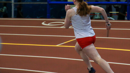 Female runner running fast on a banked indoor track holding a baton during a relay race - obrazy, fototapety, plakaty