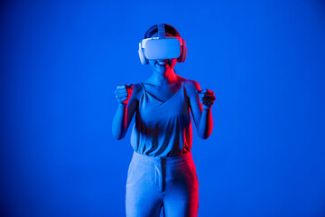 Obraz na płótnie Canvas Smart female stand surrounded by neon light wearing VR headset connecting metaverse, future cyberspace community technology. Elegant woman enjoy playing car racing games in meta world. Hallucination.
