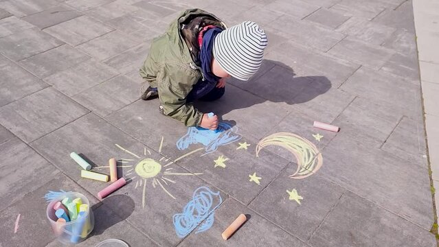 A 4-year-old boy plays on the playground in the spring, draws the sun and clouds, a month and stars on the asphalt with chalk.
