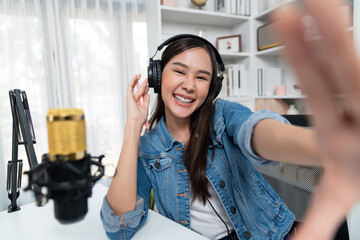 Host channel young beautiful Asian girl making selfie with smartphone wearing headphones at modern...