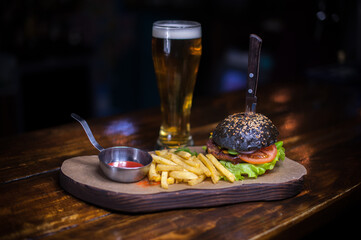 A burger with a knife sticking out of it and a glass of beer. The burger is black and has a lot of toppings. The fries are on a plate next to the burger