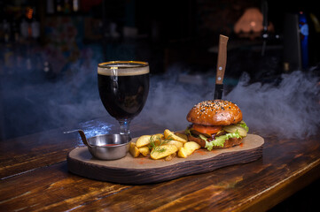A burger and fries are on a wooden table with a glass of beer. The burger is topped with lettuce and tomato, and the fries are seasoned.