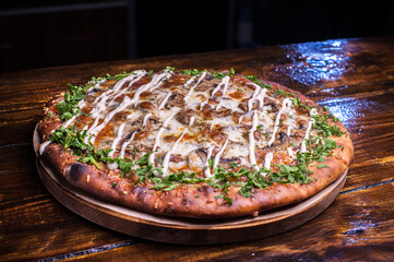 A pizza with cheese and herbs sits on a wooden table. The pizza is topped with a green herb garnish and has a creamy sauce