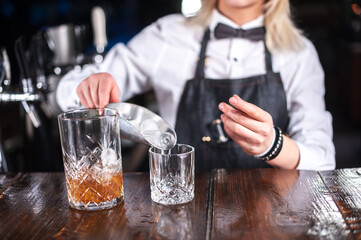 A woman is pouring a drink into a glass. The drink is a cocktail and the glass is on a wooden table
