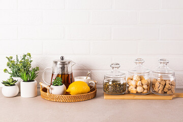 Stylish kitchen background with glass tea pot, lemon, cup, tea jars, sugar, meringue pastry. Green plants in pots. Front view.