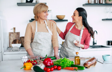 Smiling mature mother and her expressive lovely adult daughter cutting vegetables for a vegan salad together. Mid-adult woman and cute girl are preparing proper healthy meal on domestic kitchen - 777365553