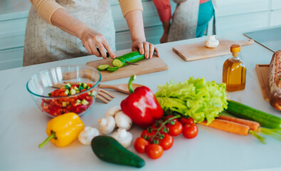 Close up image of female hands with knife cutting fresh cucumber on the table on domestic kitchen. Many ripe, tasty and colorful vegetables with olive oil. Vegan meal, healthy nutrition
