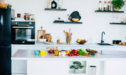 Cooking of healthy meal on domestic kitchen. Modern new light interior of kitchen with white furniture and kitchen island with many fresh vegetables on table