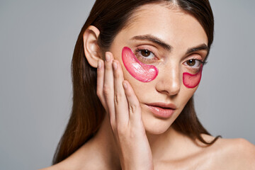 A young Caucasian woman with brunette hair wearing vibrant pink eye patches on her face, creating a...