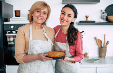 Tasty mom apple pie. Happy beautiful mature female with her attractive daughter in aprons holds and shows vegan cake after it baked on domestic kitchen on the camera. - 777366109