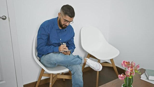 A focused bearded man sits writing in a modern room with flowers and white furniture.