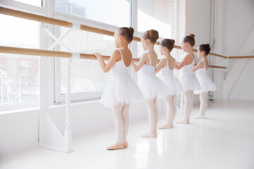 Group of children, school-girls in white dresses training poses near barre in light room. Classical...