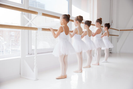Group of children, school-girls in white dresses training poses near barre in light room. Classical ballet school. Concept of art, sport, education, hobby, active lifestyle, leisure time.