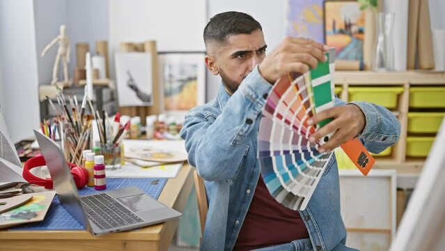A focused man with a beard examines color swatches in a creative studio environment, surrounded by paint and art supplies.